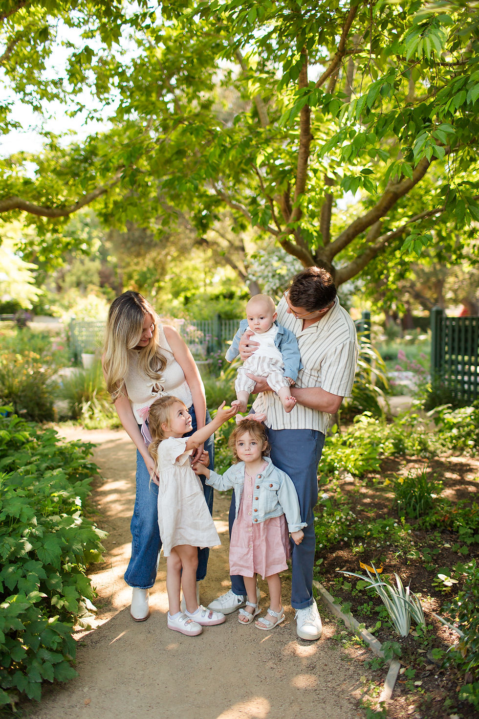 Family of five standing on a garden path under leafy trees, parents holding a baby while two young girls reach and smile. Spring Family Photoshoot at Elizabeth Gamble Garden, Palo Alto by Bay Area Photographers HKCreate