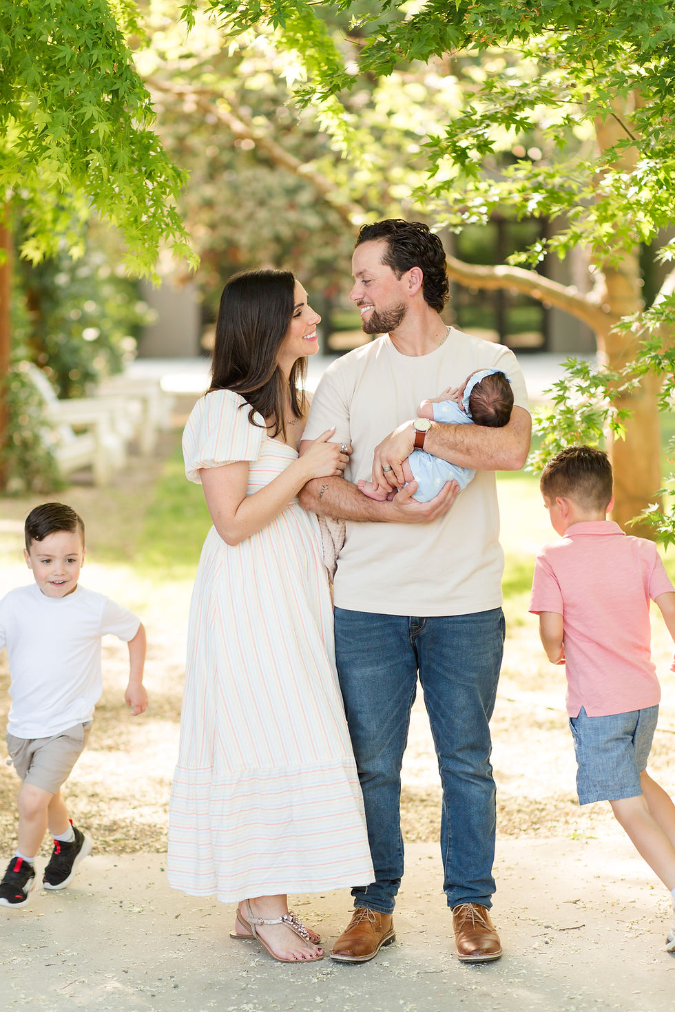 Smiling family under green trees, parents holding a baby while two young boys run nearby on a sunny garden path. Outdoor Newborn Family Photoshoot at Pageo Lavender Farm in Turlock California by Bay Area Photographers HKCreate