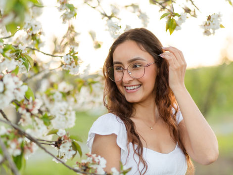 Woman smiling near blooming cherry blossoms, wearing glasses and a white dress. Sunlit orchard background, cheerful mood. High School Senior Graduation Spring Photoshoot at Saratoga Library Heritage Orchard by HKCreate Bay Area Photographers.