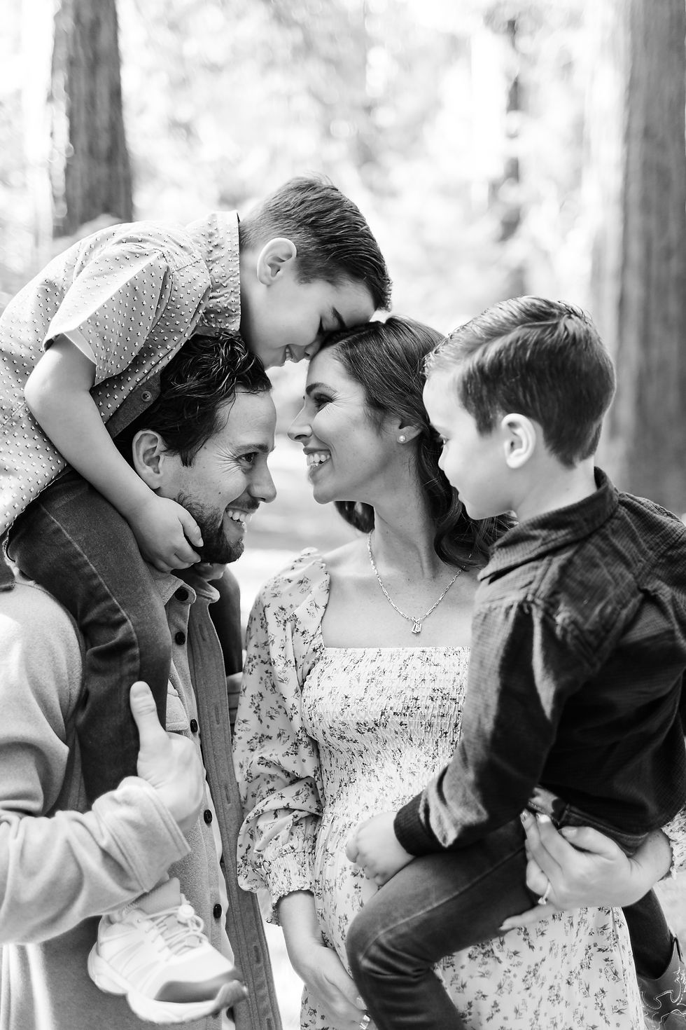 Black-and-white family portrait of smiling parents with two young boys hugging them in a wooded outdoor setting. Winter Family Maternity Photoshoot at Redwood Grove Nature Preserve, Los Altos by Bay Area Photographers HKCreate