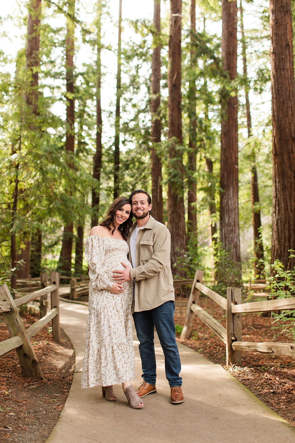 Smiling couple on a forest path, man holding pregnant woman’s belly; tall redwoods and wooden railings in soft sunlight. Winter Family Maternity Photoshoot at Redwood Grove Nature Preserve, Los Altos by Bay Area Photographers HKCreate