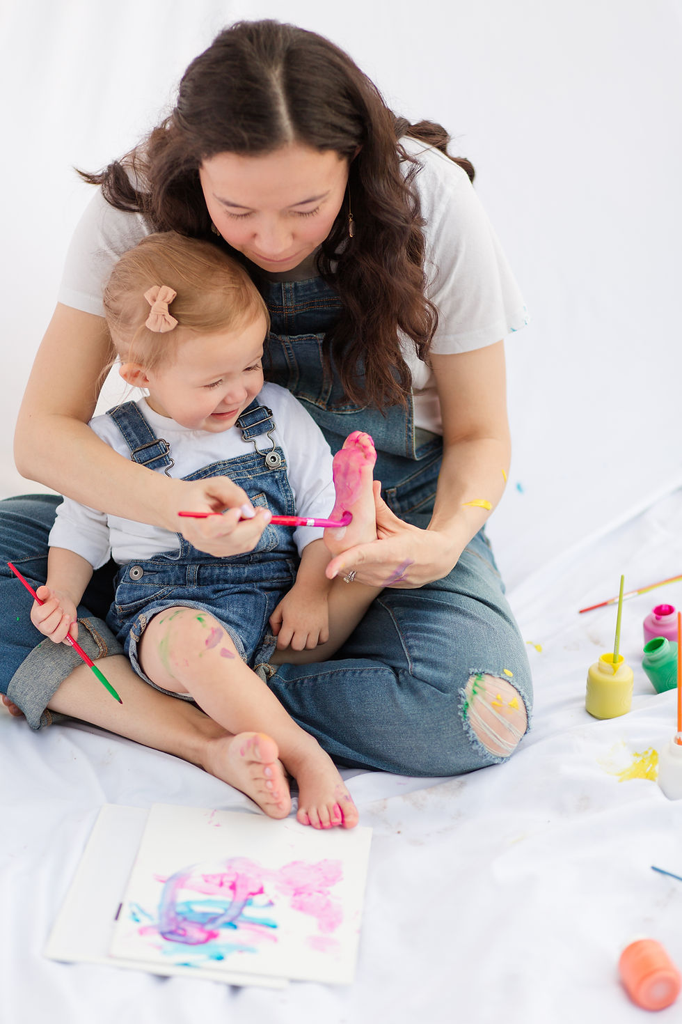 A woman and child in denim overalls paint together on paper; the child has pink paint on their foot. Bright paints surround them.
Mother and child joyfully paint each other in a playful setting. Both wear denim overalls with colorful paint on skin, against a white background.
Happy and cute and playful Mommy and Me Maternity Painting Session by HKCreate Bay Area Photographers at Guadalupe Oak Grove Park in San Jose