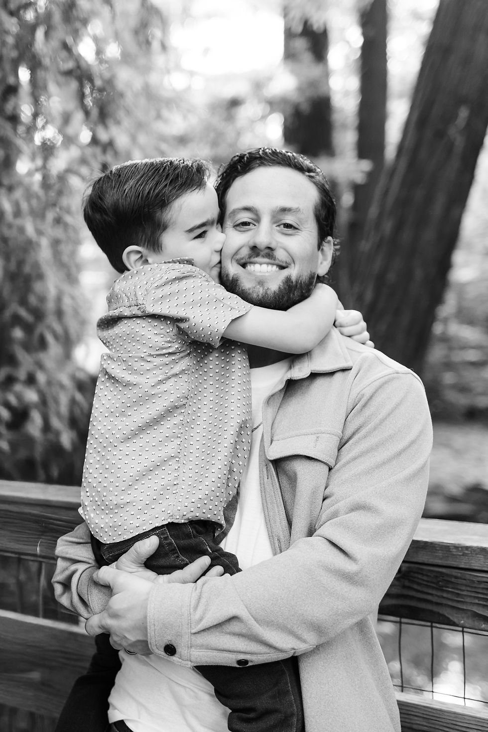 Black-and-white portrait of a smiling man holding a young boy who hugs and kisses his cheek on a wooden bridge outdoors. Winter Family Maternity Photoshoot at Redwood Grove Nature Preserve, Los Altos by Bay Area Photographers HKCreate
