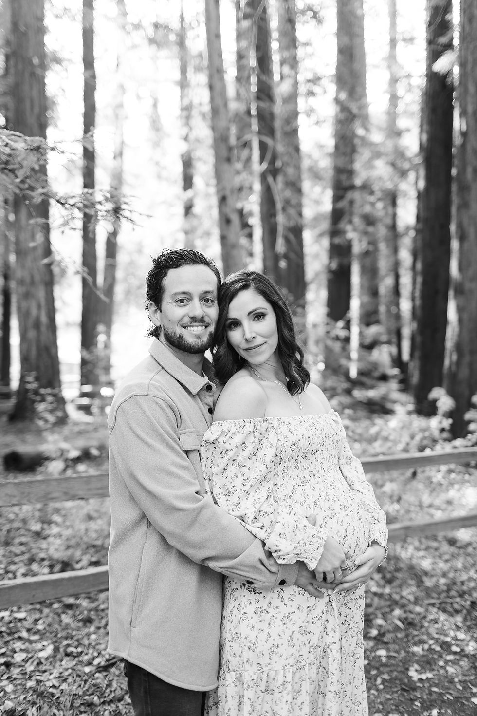 Black-and-white portrait of a smiling couple embracing in a forest; the woman wears a floral dress and cradles her pregnant belly. Winter Family Maternity Photoshoot at Redwood Grove Nature Preserve, Los Altos by Bay Area Photographers HKCreate