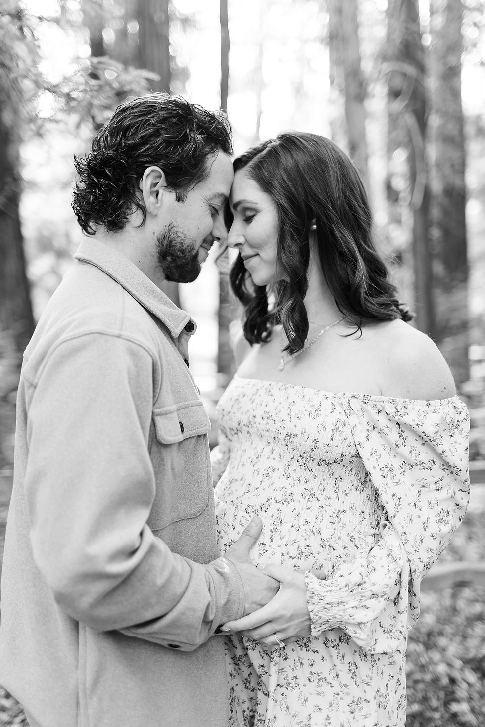 Black-and-white portrait of a couple touching foreheads in a forest, smiling softly as he holds her pregnant belly. Winter Family Maternity Photoshoot at Redwood Grove Nature Preserve, Los Altos by Bay Area Photographers HKCreate