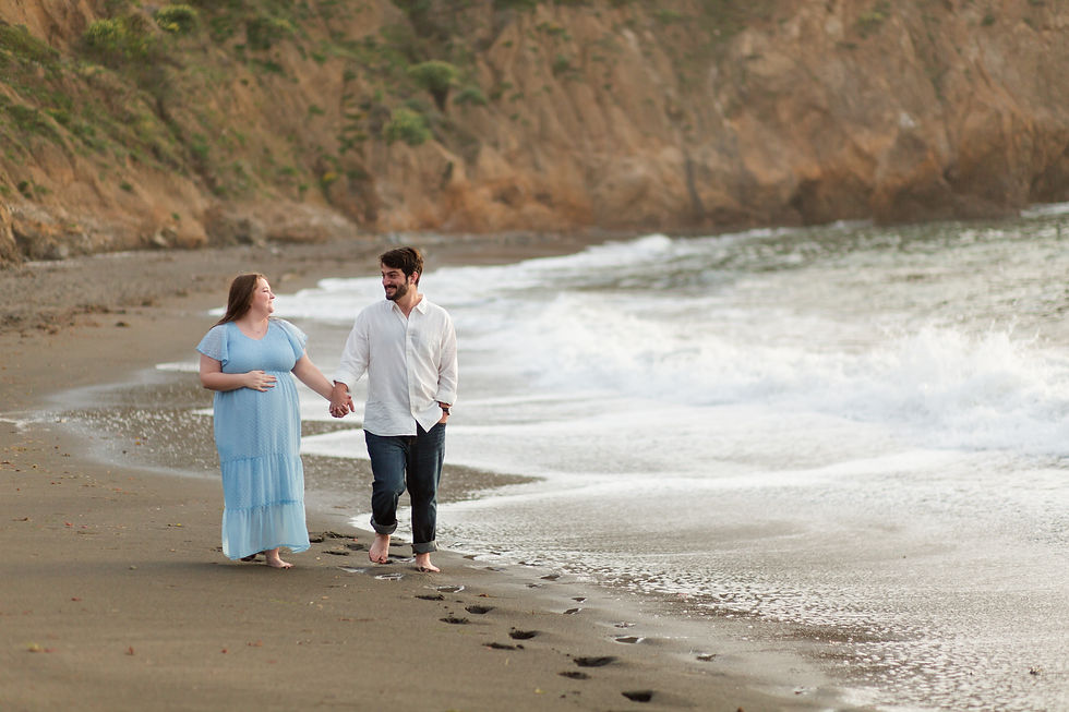 A couple walks hand in hand on a sandy beach, barefoot. The man smiles in white, and the pregnant  woman wears a blue dress. Waves crash nearby.
Rockaway Beach, Pacifica spring maternity photoshoot by Bay Area Photographers HKCreate