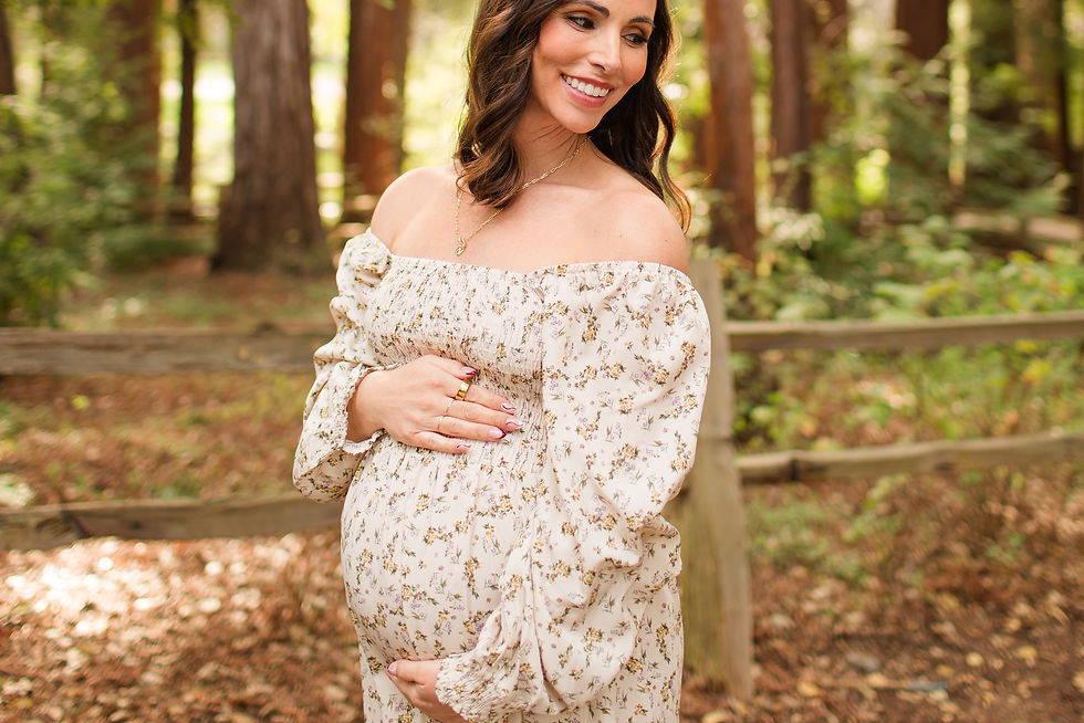 Smiling pregnant woman in a floral off-shoulder dress holds her belly in a sunlit forest with a wooden fence. Winter Family Maternity Photoshoot at Redwood Grove Nature Preserve, Los Altos by Bay Area Photographers HKCreate