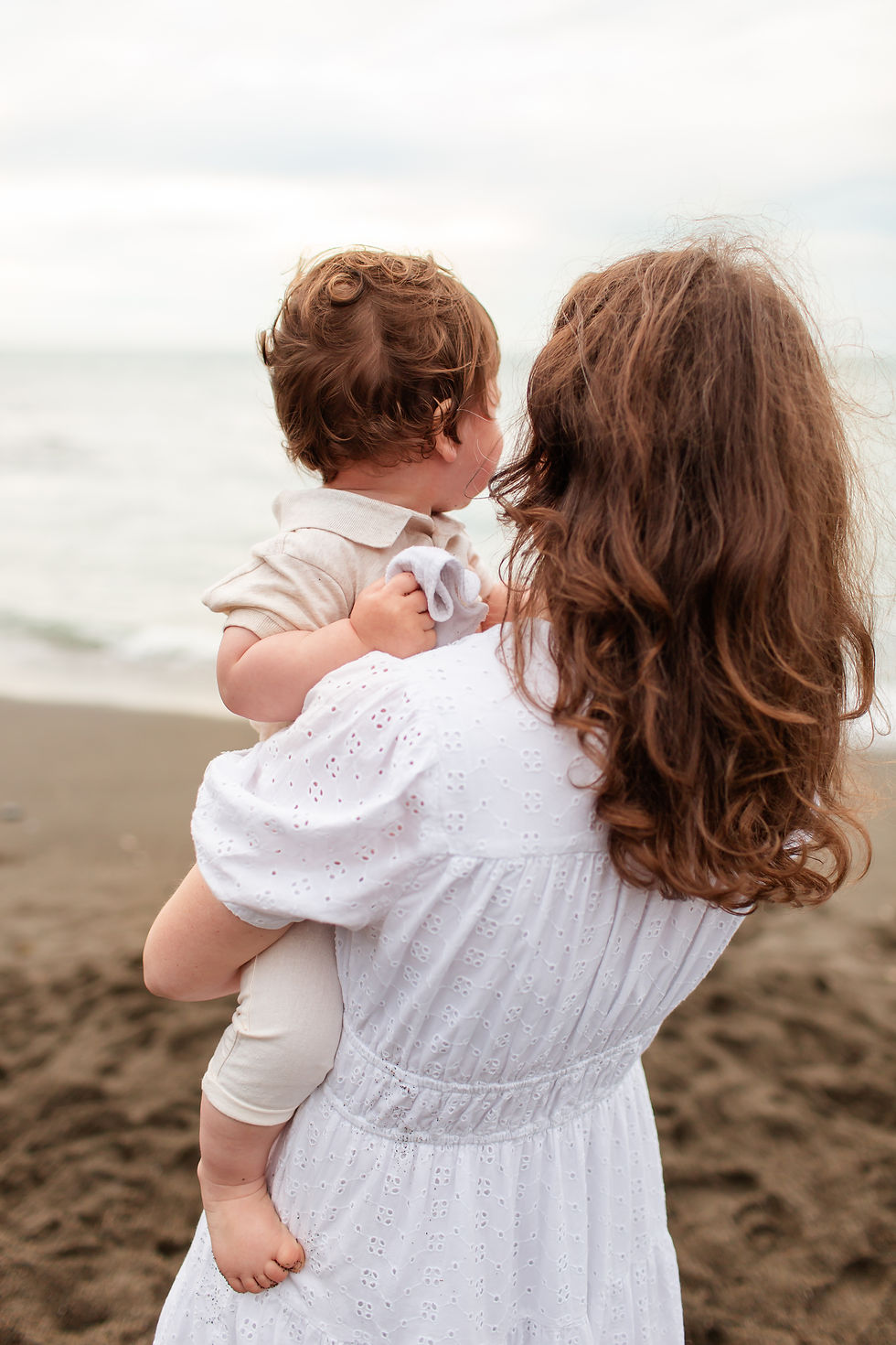 Mother in a white dress holds a toddler on a cloudy beach, both facing the calm sea. Overcast Beach Family Photoshoot at Rockaway Beach in Pacifica by Bay Area Photographers HKCreate