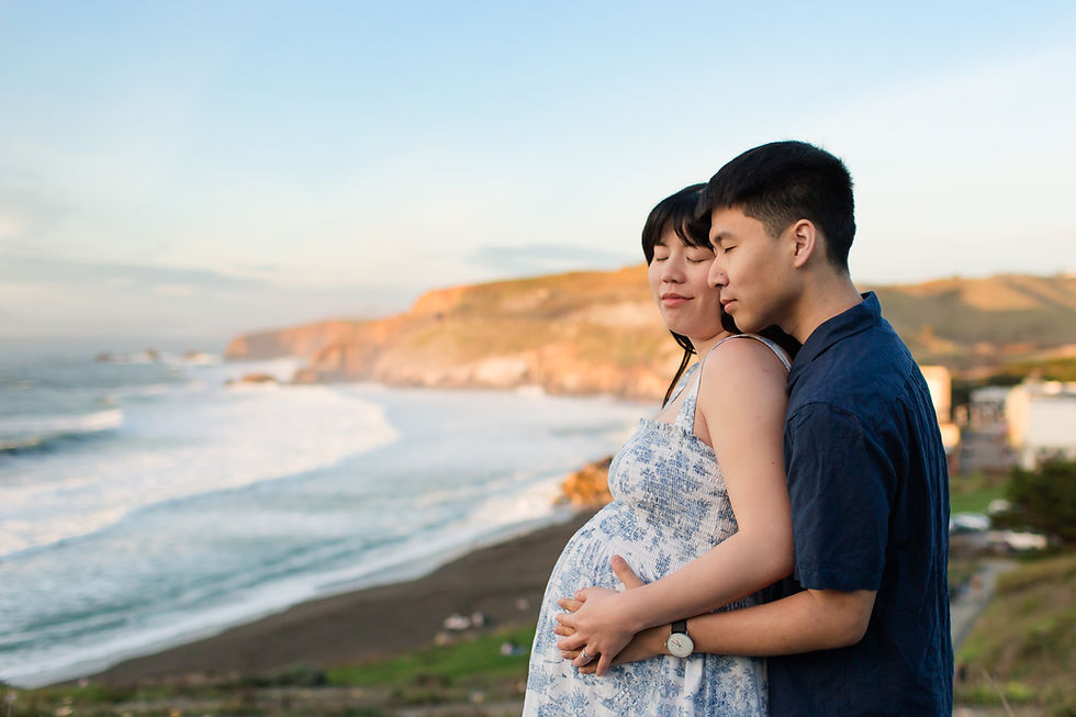 A couple embraces on a scenic cliffside at sunset, with waves crashing below. Both appear serene and content, against a vivid coastal backdrop.
Sunset Spring Beach Maternity Session at Rockaway Beach in Pacifica by HKCreate Bay Area Photographers