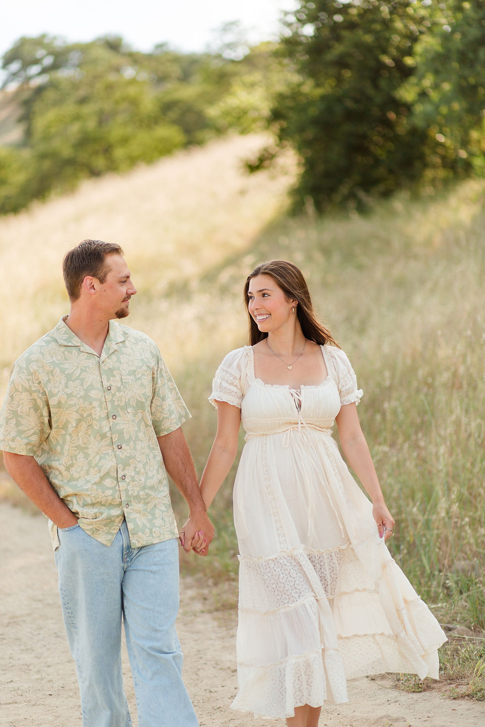 A couple holds hands, walking on a grassy path. The man wears a floral shirt; the woman in a white free people dress. They smile at each other, sunny day.
Spring family photoshoot at Guadalupe Oak Grove Park in San Jose by HKCreate Bay Area Photographers.