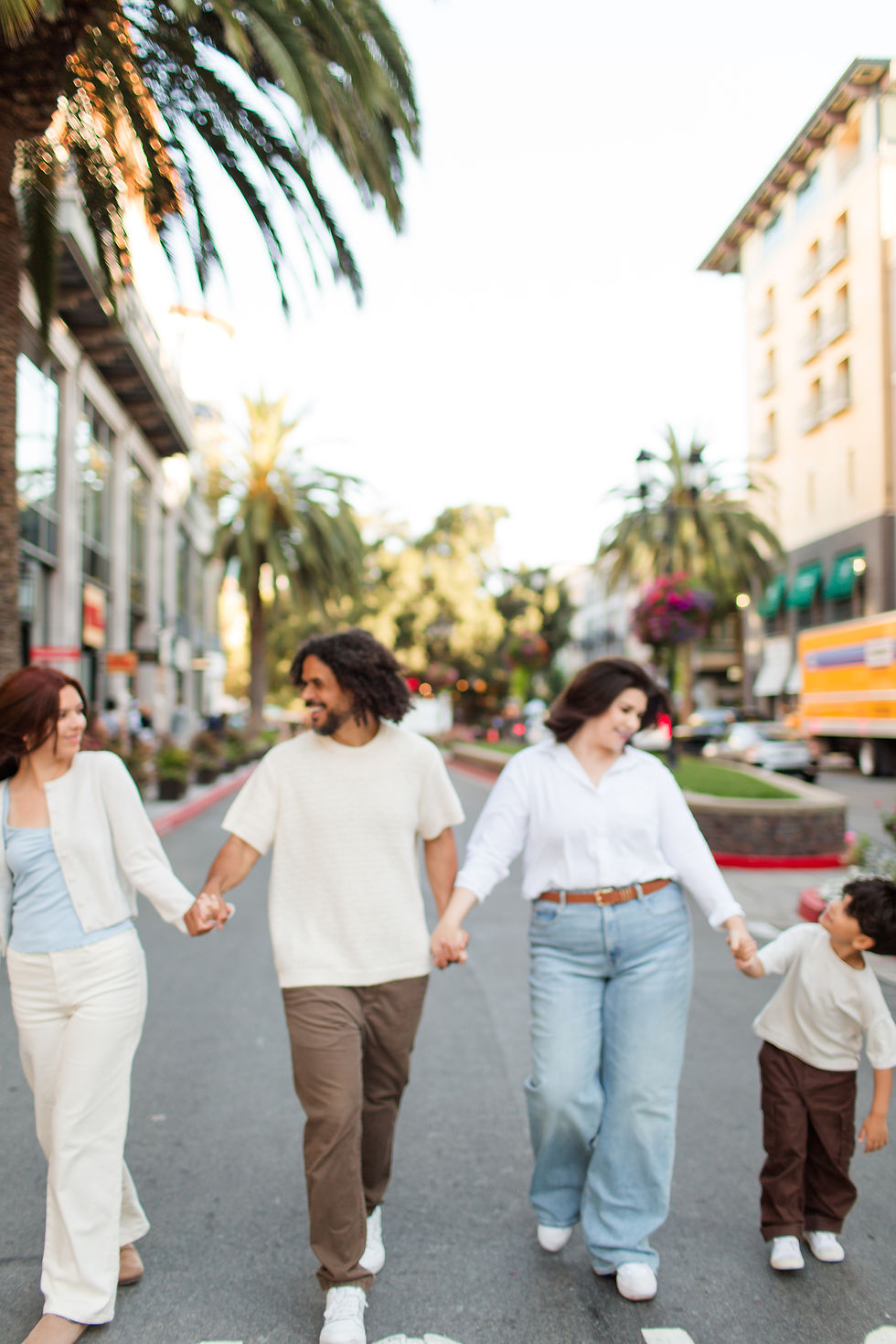 Four people walk hand in hand down a street lined with palm trees. They are smiling, and the mood is joyful. Buildings are visible in the background.
Santana Row Shopping Center, San Jose Urban Family Summer Photoshoot by HKCreate Bay Area Family Photographers HKCreate
