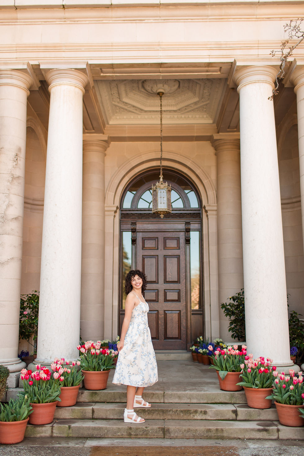 Smiling woman in a white floral dress stands on stone steps between columns, with potted tulips and a dark wooden door behind her.
Spring High School Grad Photoshoot at Filoli Historic House and Gardens, Woodside by Bay Area Photographers HKCreate