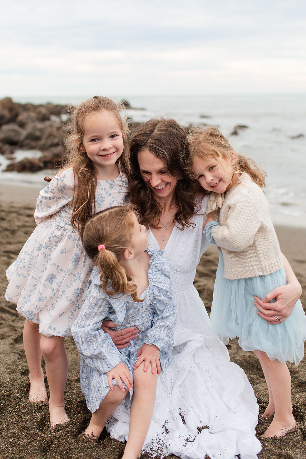 Smiling mother hugs three young daughters on a rocky beach, all in light blue and white dresses with the sea behind them. Overcast Beach Family Photoshoot at Rockaway Beach in Pacifica by Bay Area Photographers HKCreate