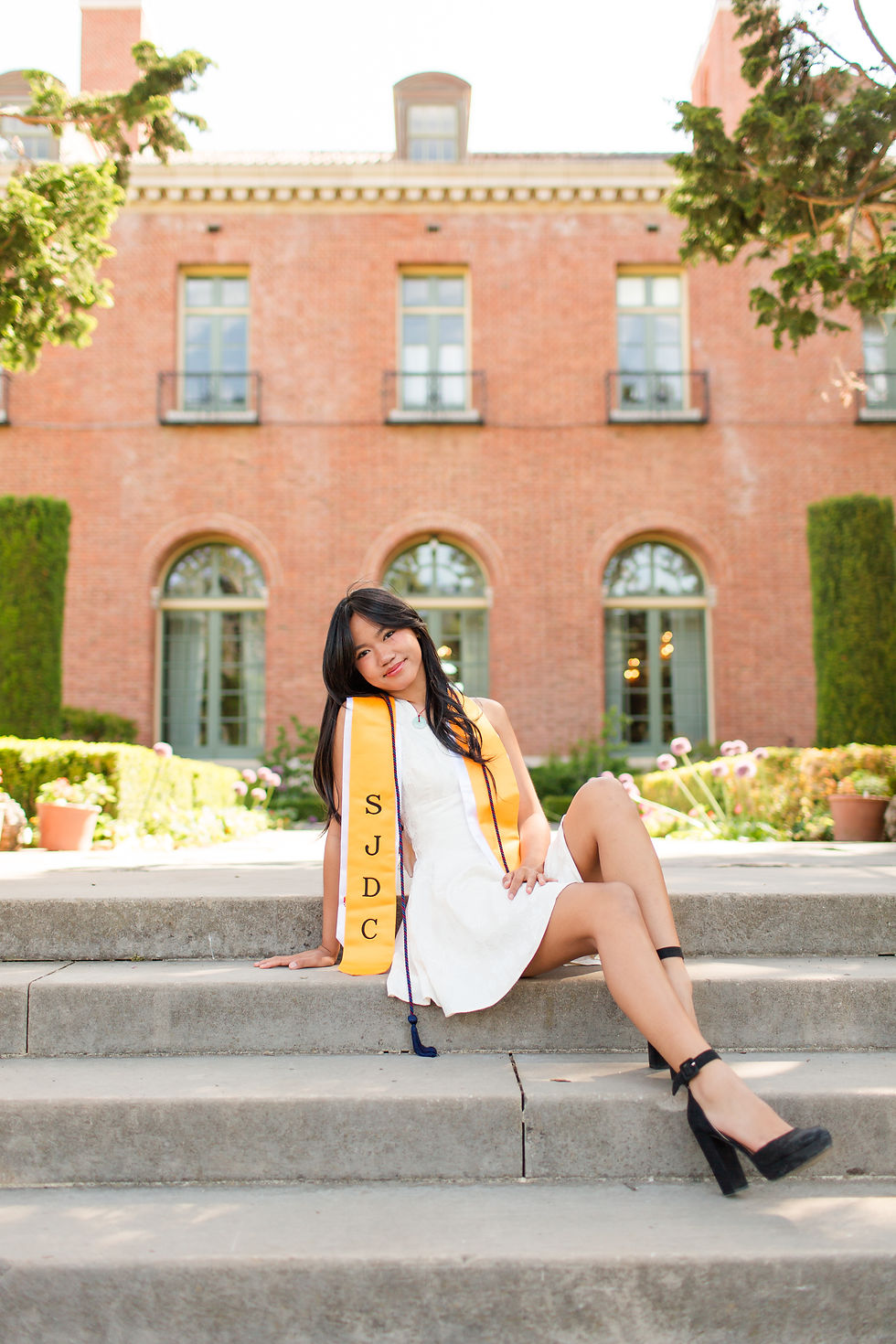 Young woman in white dress and yellow sash sits on steps before a brick building, smiling outdoors. spring high school college graduation photoshoot sisters at filoli house and garden, woodside by bay area photographers hkcreate