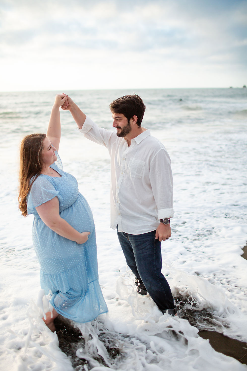 Pregnant woman in blue dress and man in white shirt dance playfully on the beach, waves hitting their feet under a cloudy sky.
Rockaway Beach, Pacifica spring maternity photoshoot by Bay Area Photographers HKCreate