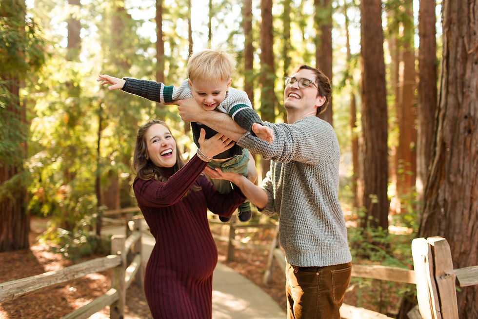A joyful family plays in a sunny forest. Parents lift a smiling toddler; the mom wears a burgundy dress, and the dad a gray sweater.
Winter Family Session at Redwood Grove Nature Preserve in Los Altos by Bay Area Photographers HKCreate