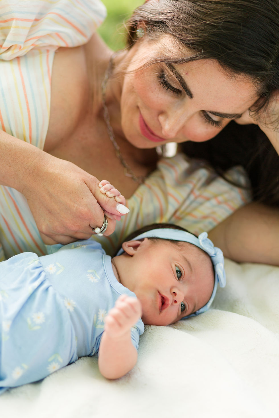 Mother smiling over newborn baby in a blue outfit and headband, lying on a white blanket in soft daylight. Outdoor Newborn Family Photoshoot at Pageo Lavender Farm in Turlock California by Bay Area Photographers HKCreate
