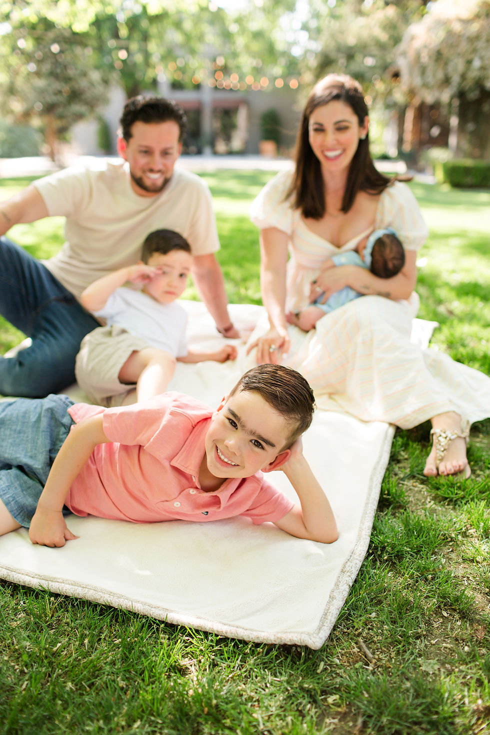 Smiling family of five relaxing on a white blanket on a sunny lawn, with a mother holding a baby and two boys in the foreground. Outdoor Newborn Family Photoshoot at Pageo Lavender Farm in Turlock California by Bay Area Photographers HKCreate