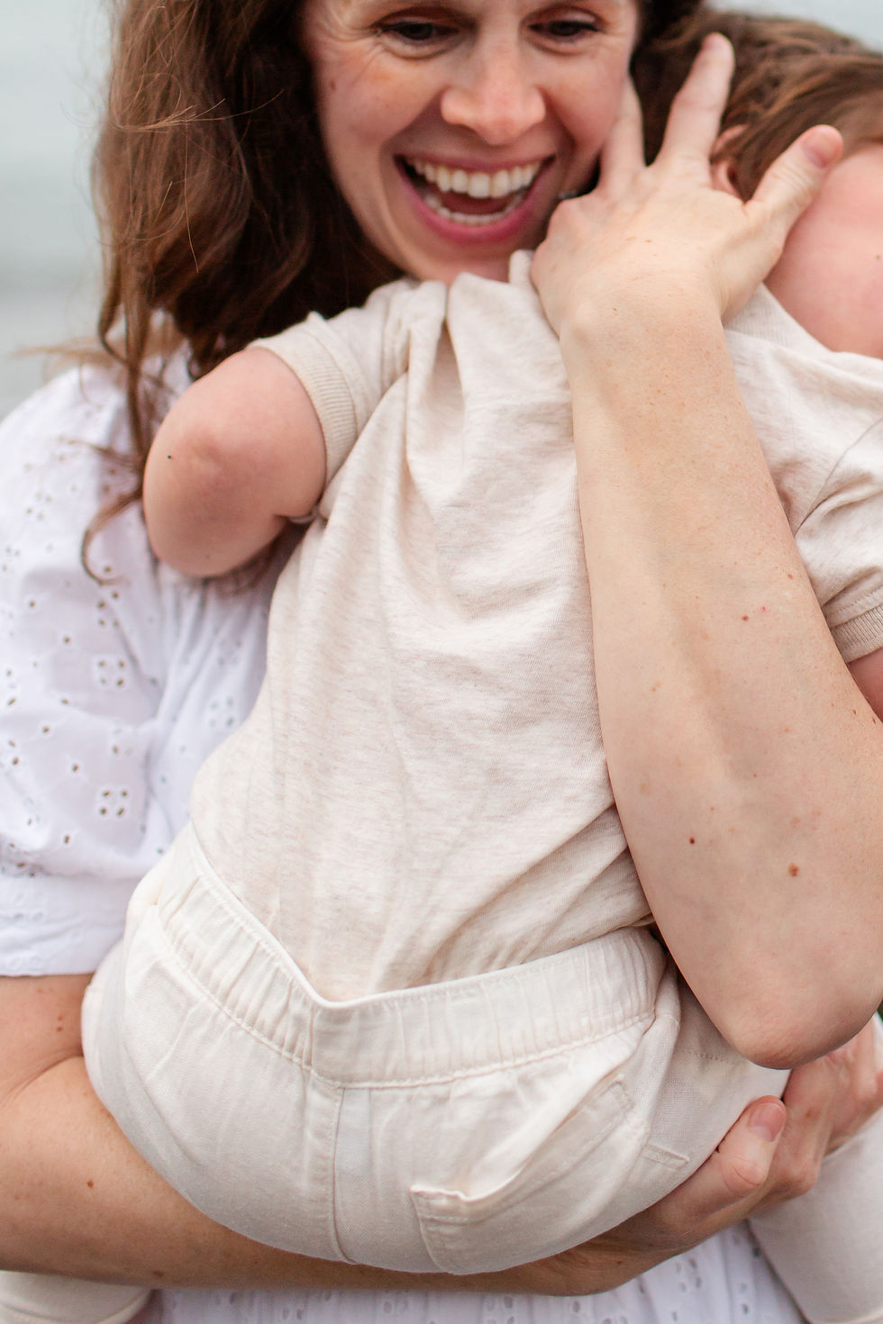 Smiling mom hugs a baby in a cream outfit close to her face, conveying joy and affection against a soft, bright background. Overcast Beach Family Photoshoot at Rockaway Beach in Pacifica by Bay Area Photographers HKCreate