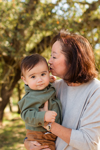 Grandma holding grandson kissing the side of his head. Warm and happy mood. One year old birthday boy legacy fall family photoshoot at Guadalupe Oak Grove Park, San Jose by HKCreate Bay Area Photographers.