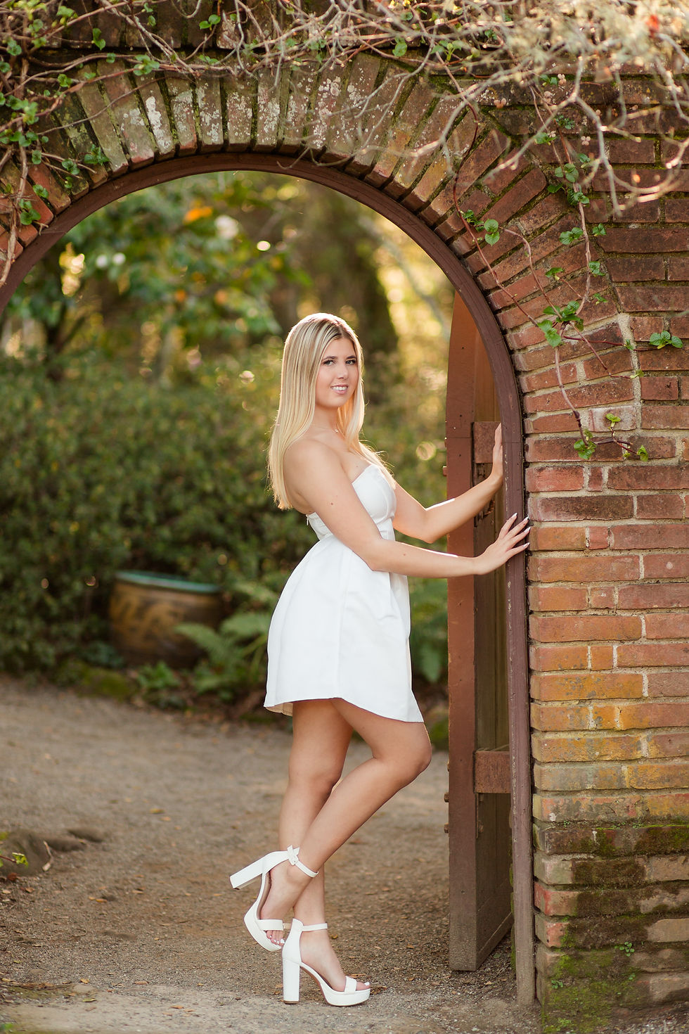 Woman in a white dress and heels stands under a brick archway, smiling, in a lush outdoor garden setting with green foliage.
Spring High School Graduation Photoshoot at Filoli Historic House and Garden by Bay Area San Jose Photographers HKCreate