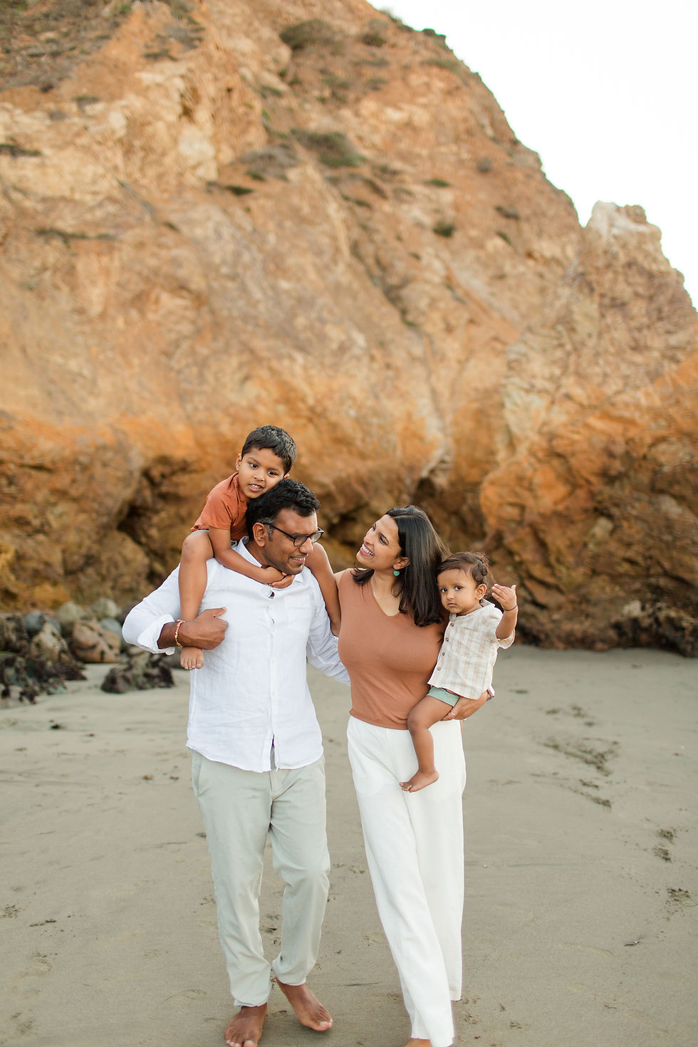 Family of four walking on a sandy beach, father carrying a child on shoulders. Rocky cliffs in the background, warm and cheerful mood.
Rockaway Beach Pacifica Fun Playful and Cute Family Photoshoot by HKCreate Bay Area Photographers.