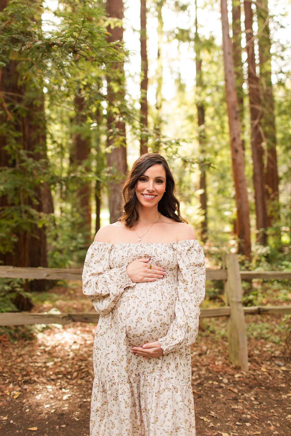 Smiling pregnant woman in a floral off-shoulder dress stands in a sunlit forest, hands on her belly. Winter Family Maternity Photoshoot at Redwood Grove Nature Preserve, Los Altos by Bay Area Photographers HKCreate