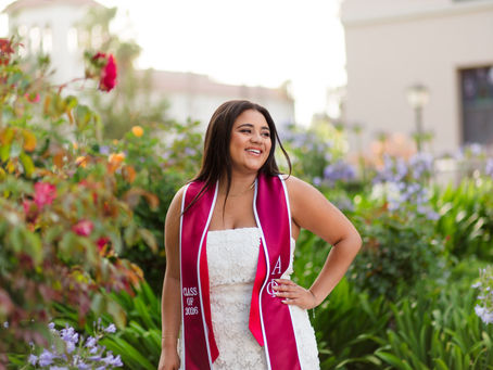 Smiling young woman in a white dress and pink Class of 2026 sash stands in a flower garden, hand on hip. Santa Clara University Grad Photoshoot by Bay Area Photographers HKCreate