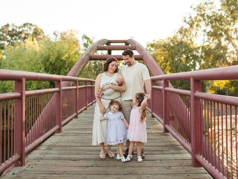 Family of five on a red bridge. Parents hold a newborn baby; two girls stand beside them. Green trees in the background, creating a serene mood. Summer Outdoor Newborn Photoshoot at Vasona Lake Park, Los Gatos by HKCreate Bay Area Photographers
