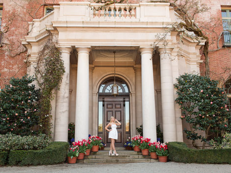Teenage girl in white dress poses on steps of grand entrance with columns, surrounded by potted tulips, against a brick building backdrop. About the Location Guide for Filoli Historic House and Garden, Woodside California by HKCreate Bay Area Photographers