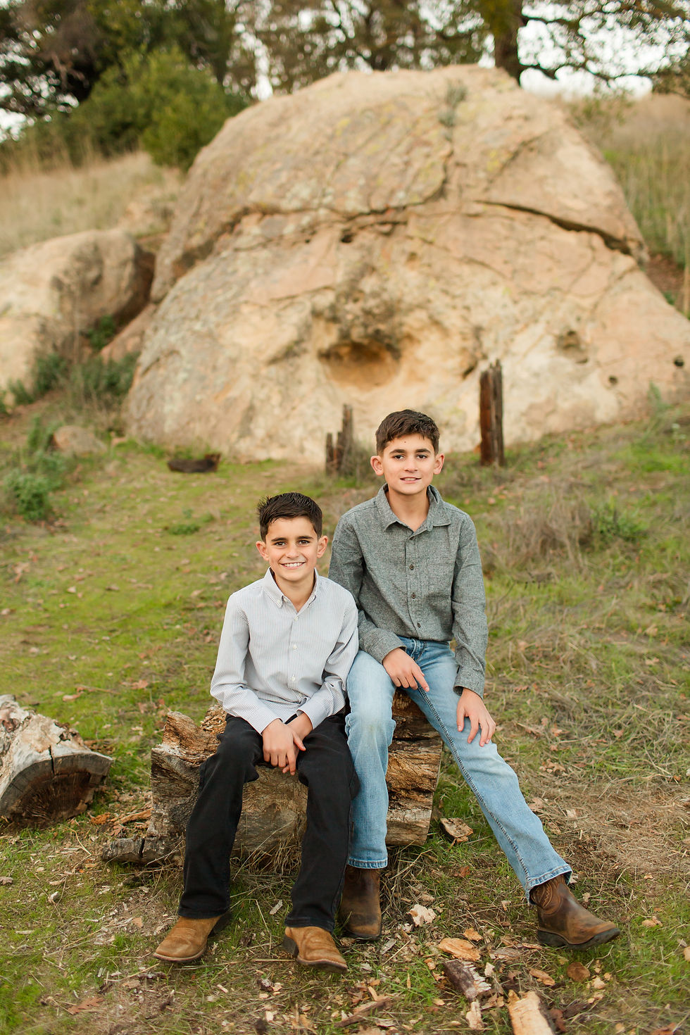 Two boys sit on logs in front of a large rock in a grassy area, smiling. The younger one wears a striped shirt; the older one wears jeans.
Sunset fall family photoshoot at Guadalupe Oak Grove Park in San Jose by Bay Area Photographers HKCreate