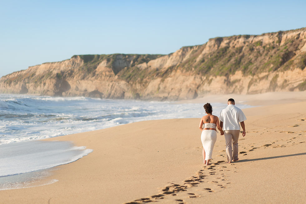 Couple walking hand in hand on a sandy beach, leaving footprints. Cliffs and ocean waves in the background under a clear blue sky. hkcreate bay area photographers half moon bay california beach maternity session sunset golden hour