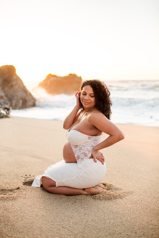 Smiling woman in white dress kneels on sandy beach at sunset. Waves crash behind her. Joyful and serene mood. Maternity Session at Cowell Ranch State Beach in Half Moon Bay by HKCreate Bay Area Photographers