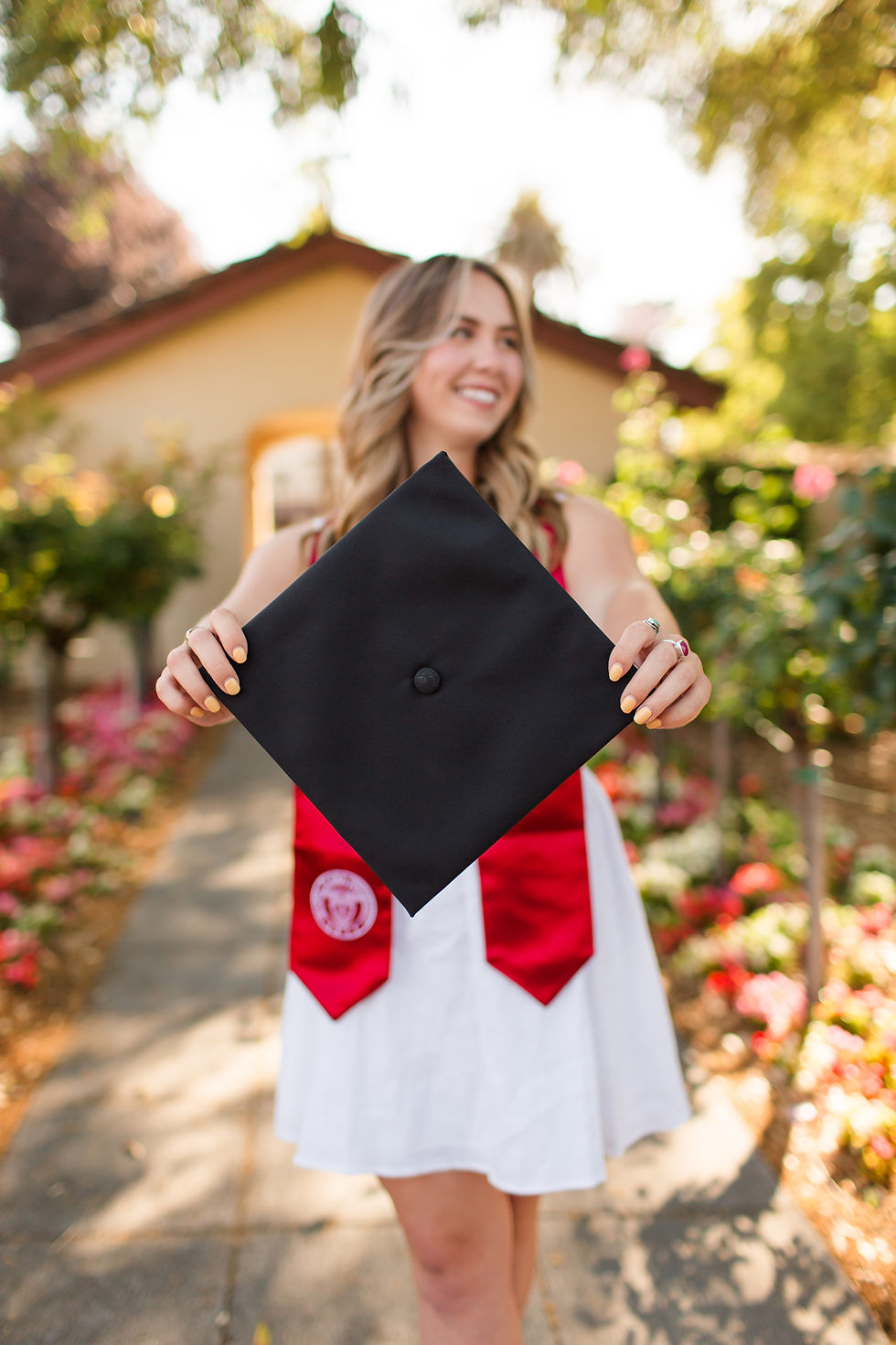 Graduate in white dress and red stole holds cap in garden, smiling. Background of flowers and house suggests joyful celebration.
Santa Clara University graduation photoshoot at the Mission Gardens by Bay Area Photographers HKCreate.