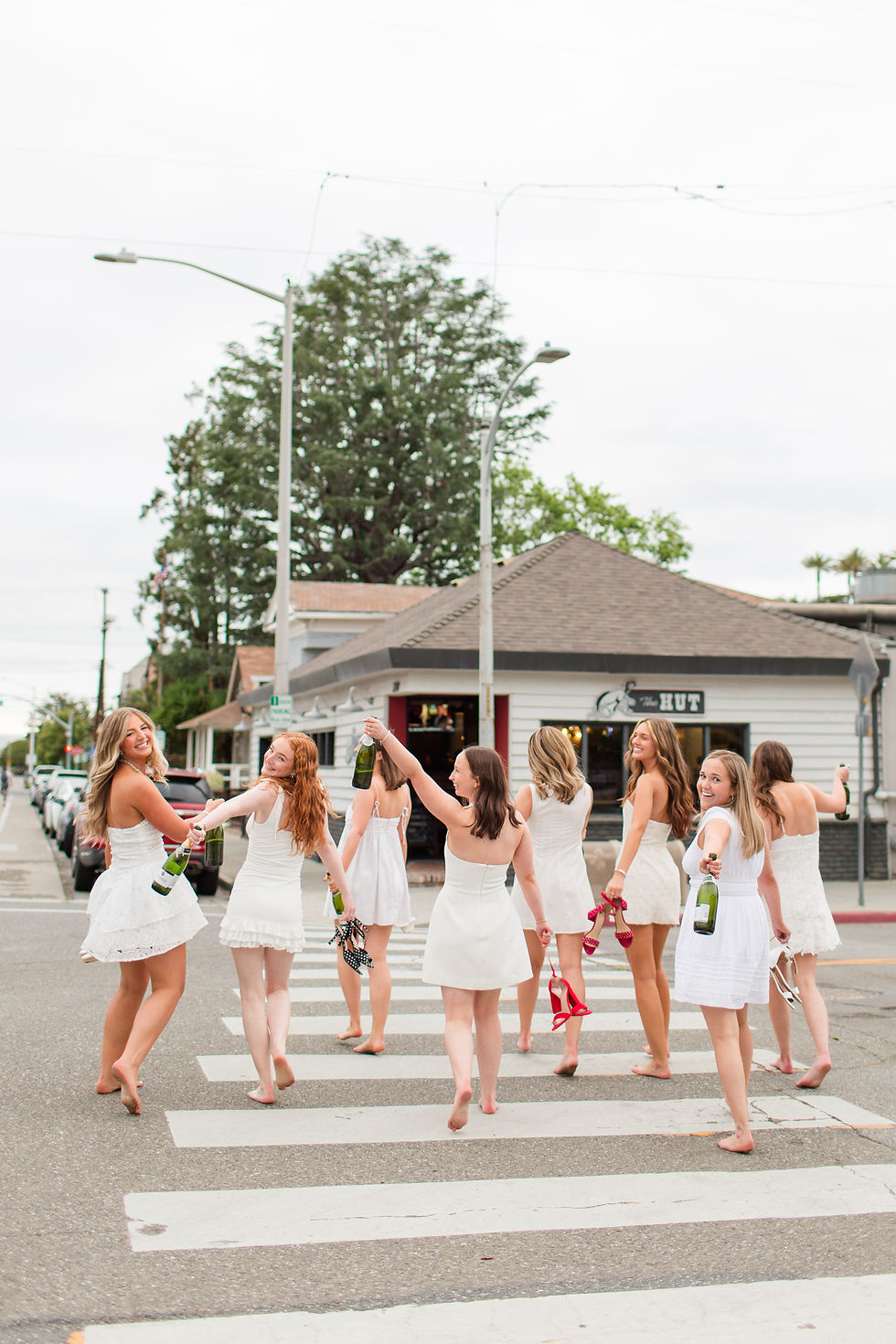 Eight women in white dresses walk barefoot across a crosswalk, holding shoes and champagne bottles in front of The Hut campus restaurant. Santa Clara University fun group grad photoshoot by bay area photographers HKCreate