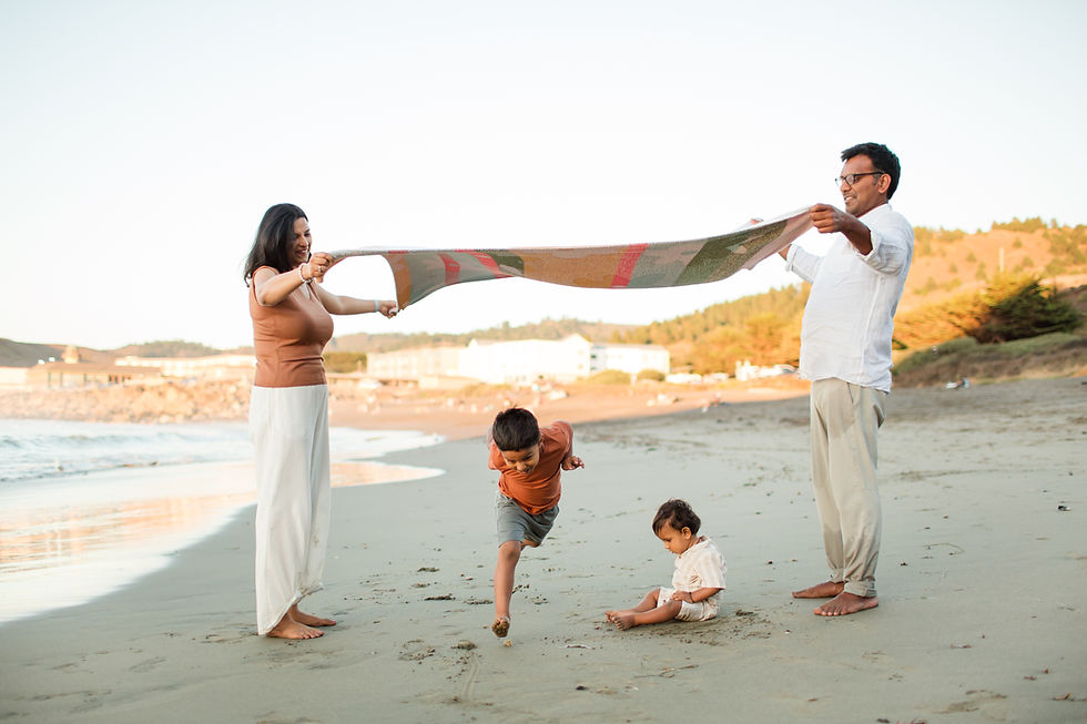 Family at beach with parents holding a colorful blanket. Child jumps joyfully beneath, while a toddler sits on the sand. Clear sky.
Rockaway Beach Pacifica Fun Playful and Cute Family Photoshoot by HKCreate Bay Area Photographers.