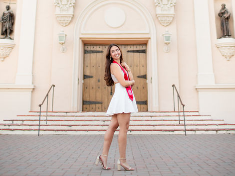 Smiling girl wearing short white dress and red stole in front of the Santa Clara University mission on campus. Santa Clara University Grad Photoshoot by Bay Area Photographers HKCreate