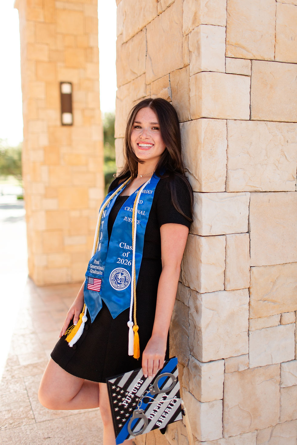 Smiling graduate in black dress leans on a stone pillar, wearing a blue Criminal Justice stole labeled Class of 2026 and holding a decorated cap.
College Grad photoshoot at Sonoma State University  by Bay Area Photographers HKCreate