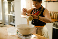 A woman wearing a black tank top, pouring sourdough starter into a bowl on a scale, in a brightly lit kitchen setting.