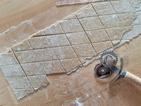 A sheet of whole grain pasta dough on a wooden counter top with a pastry cutter beside it. The pasta dough is cut into diamonds.