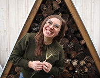A woman wearing a green sweater, posing for a photo in front of an a-frame structure of firewood. She has a big smile on her face and is holding two wheat stalks