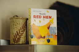 The hardcover book 'The Little Red Hen', with a red hen wearing a yellow rain jacket. The book is standing up beside a pottery urn with the brown imprint of a fern on it.