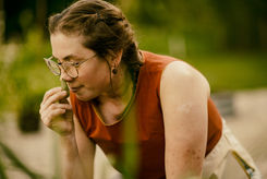 A woman with her hair braided, wearing a red and white shirt, bending down in the garden to smell the lemon balm.