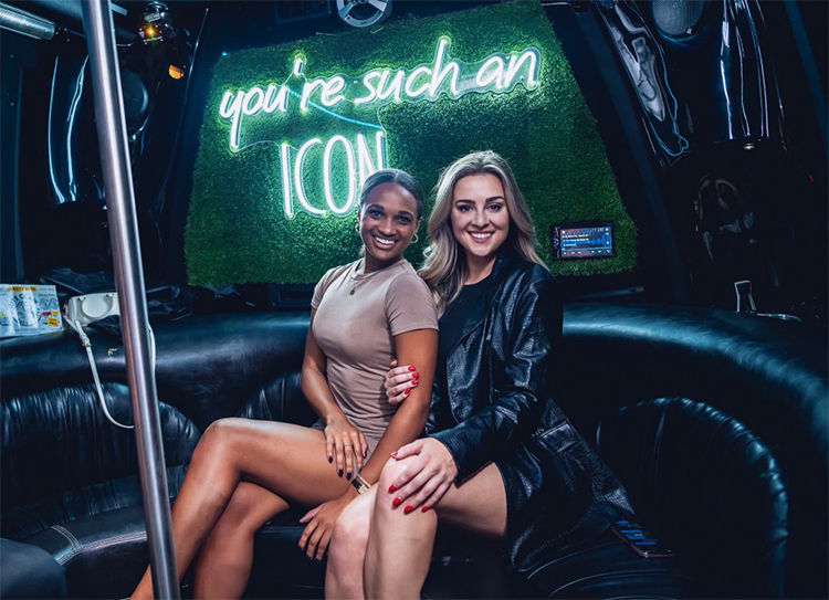 Two women smiling in a party bus, with a neon sign reading 'you're such an ICON'