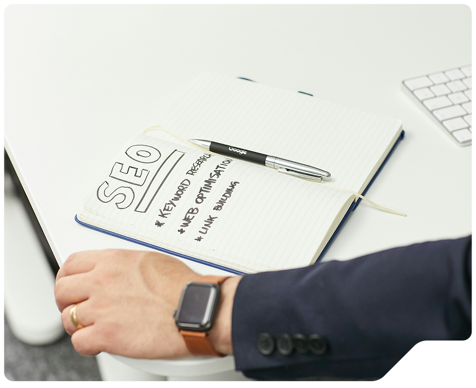 Zoek Marketing moves SEO from your notebook to your computer screen with this image of a notebook on desk, text shows an SEO checklist: "Keyword Research," "Web Optimization," "Link Building." Man's hand with smartwatch nearby.