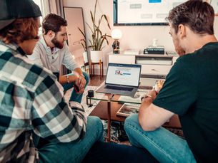 Three men casually dressed huddle around a laptop in an office, studying a web page; warm colors, representing a Zoek Marketing client.