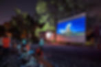 People watch a movie outdoors at night with a castle on screen. Trees surround the area. Some seated on benches, others on blankets. Relaxed mood.