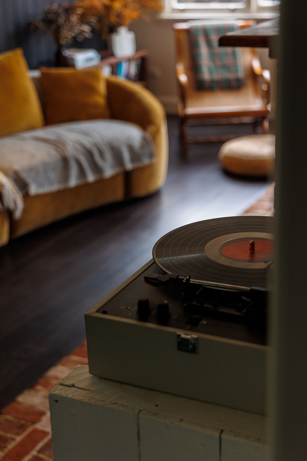 Vinyl record player in a cozy living room with a mustard sofa, chair, and soft warm light.