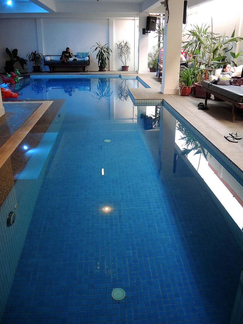 Indoor pool area with blue tiles at Siem Reap Hostel. Person reading on left sofa, another resting on right sofa. Plants and colorful inflatables add vibrant touch.