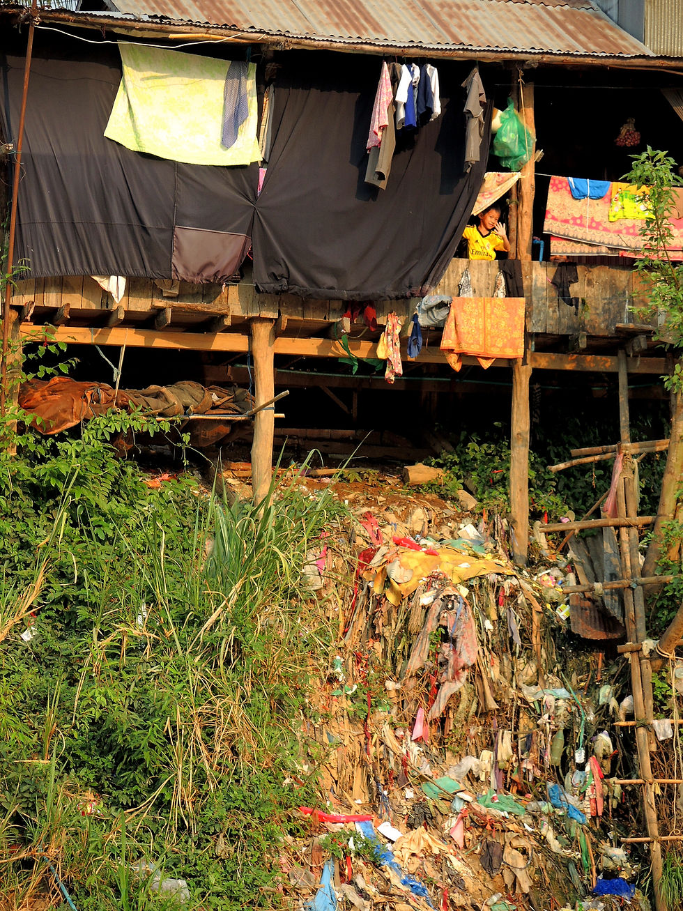 A makeshift house on stilts with hanging clothes, surrounded by greenery and trash. A person in a yellow shirt peers out. Rustic mood.